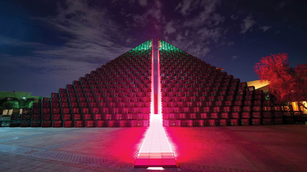 Rashed AlShashai’s installation The Fifth Pyramid (2024), a monumental green pyramid constructed from stacked petrochemical pallets, split down the middle to reveal a glowing fuchsia-lit path, shown illuminated at night at the King Abdulaziz Historical Center in Riyadh.
