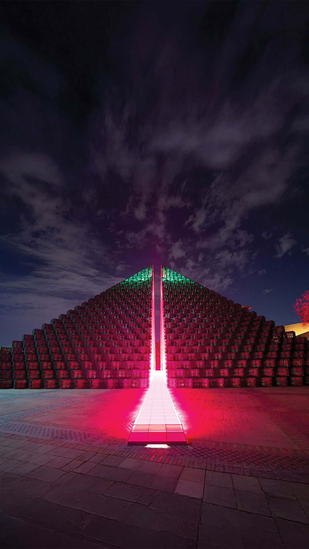 Rashed AlShashai’s installation The Fifth Pyramid (2024), a monumental green pyramid constructed from stacked petrochemical pallets, split down the middle to reveal a glowing fuchsia-lit path, shown illuminated at night at the King Abdulaziz Historical Center in Riyadh.